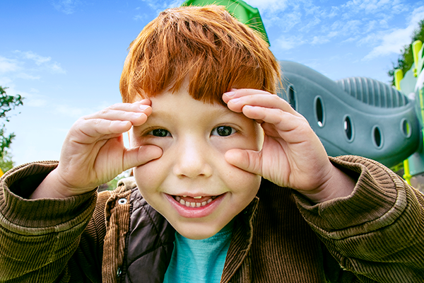 Red-headed boy looking closely at the camera with his hands around his eyes.